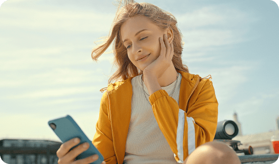 Mulher jovem, sentada, a sorrir para o telemóvel que segura na mão